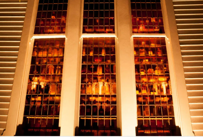 Three tall leaded stained-glass window panels glowing warm amber behind white mullions, flanked by louvered shutters.