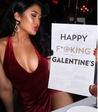 Glamorous woman in a red velvet halter dress and sparkly earrings puckering for a photo while holding a menu-style sign that reads "HAPPY F*CKING GALENTINE'S" in a dimly lit restaurant.