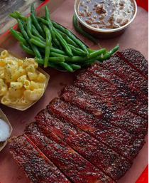 Sliced smoked barbecue brisket with peppery bark on a platter, served with creamy mac and cheese, tender green beans and a cup of saucy baked beans.
