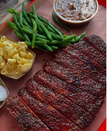 Sliced smoked barbecue brisket with peppery bark on a platter, served with creamy mac and cheese, tender green beans and a cup of saucy baked beans.