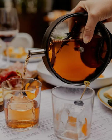 Hand pouring an amber craft cocktail from a small teapot into a rocks glass with a large ice cube and orange peel garnish on a dining table with plates and charcuterie.