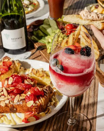 Sunlit brunch table with a frosty berry cocktail in a wine glass topped with blueberries and blackberries, almond-topped strawberry toast on a plate, grilled bread and avocado on a wooden board, and a wine bottle nearby.