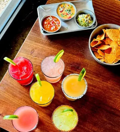 Overhead view of seven colorful cocktails with lime garnishes on a wooden restaurant table by a window, alongside a bowl of tortilla chips and three small bowls of salsa and guacamole.