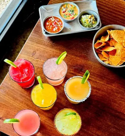 Overhead view of seven colorful cocktails with lime garnishes on a wooden restaurant table by a window, alongside a bowl of tortilla chips and three small bowls of salsa and guacamole.