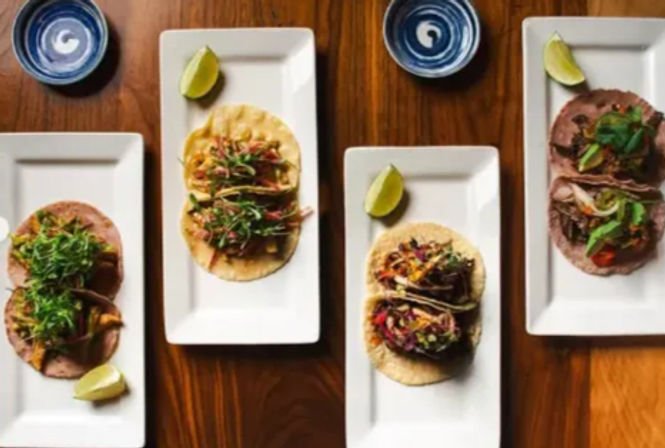 Four white rectangular plates of gourmet tacos with colorful toppings and lime wedges on a wooden table, plus two small blue dipping bowls
