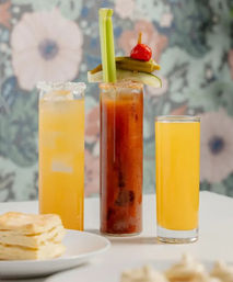 Three tall brunch cocktails on a white table: a Bloody Mary with celery stalk, pickle and cherry pepper garnish flanked by two bright orange mimosa-style drinks, stacked biscuits and a floral wallpaper backdrop.
