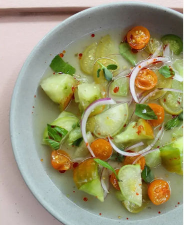 Bright summer cucumber and orange cherry tomato salad with thin red onion slices, basil microgreens, red chili flakes and citrus vinaigrette on a gray ceramic plate over a pink tabletop.