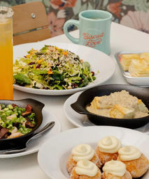 Colorful brunch spread on a cafe table: green salad with seeds and cranberries, skillet biscuits smothered in gravy, mini cream-topped doughnuts, vegetable skillet, orange juice and coffee mug.