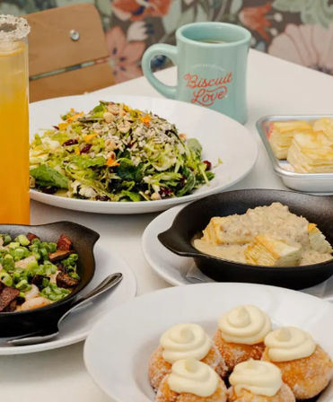 Colorful brunch spread on a cafe table: green salad with seeds and cranberries, skillet biscuits smothered in gravy, mini cream-topped doughnuts, vegetable skillet, orange juice and coffee mug.