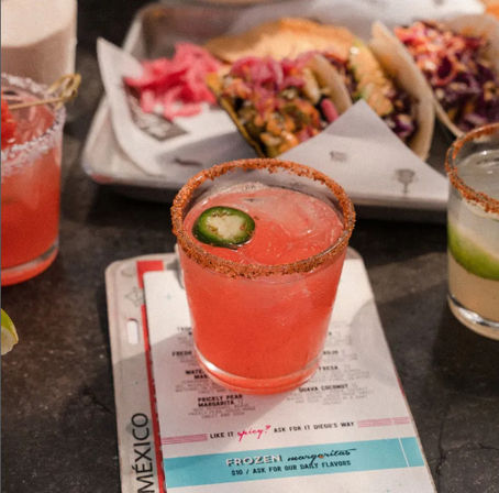 Spicy pink margarita with chili-salt rim and jalapeño slice, served on a menu beside a tray of colorful street-style tacos and drinks in a Mexican-inspired eatery