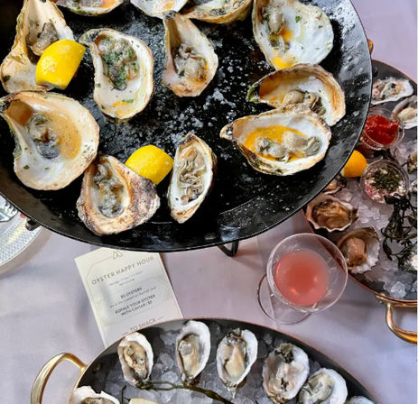 Top-down view of oysters on the half shell — grilled and raw — arranged on black platters and ice with lemon wedges and a pink cocktail for a seafood happy hour.