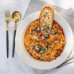 Overhead shot of Italian-inspired baked cheesy tortellini in marinara with wilted spinach and parsley, topped with a toasted garlic baguette slice on a white plate beside gold flatware and a gray napkin on a marble table.