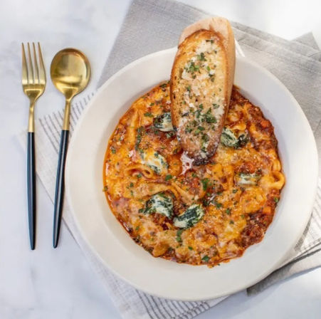 Overhead shot of Italian-inspired baked cheesy tortellini in marinara with wilted spinach and parsley, topped with a toasted garlic baguette slice on a white plate beside gold flatware and a gray napkin on a marble table.