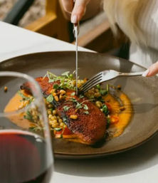 Pan-seared fish fillet with a crispy crust on a bed of lentils and orange puree, topped with microgreens and being cut with fork and knife at a restaurant table next to a glass of red wine.