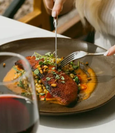 Pan-seared fish fillet with a crispy crust on a bed of lentils and orange puree, topped with microgreens and being cut with fork and knife at a restaurant table next to a glass of red wine.