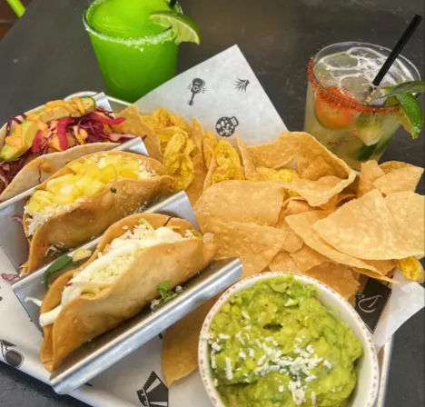 Three Mexican soft-shell tacos in a metal rack with cabbage, pineapple and crema, served with tortilla and plantain chips, a bowl of guacamole, a frozen lime margarita and a chili-rimmed cocktail.
