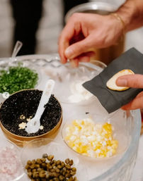 Close-up of a gourmet caviar station — hands topping a cracker with glossy black caviar, surrounded by a tin of caviar with spoon and bowls of chopped egg, capers, chives and minced shallot on a serving tray.
