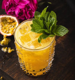 Iced passionfruit cocktail in a cut-glass tumbler topped with fresh mint, with halved passionfruit and a pink flower on a dark wooden table