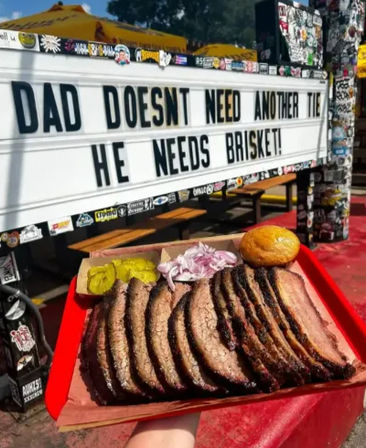 Smoked brisket slices on a red tray with pickles, sliced onions and a bun, held in front of an outdoor marquee reading 'Dad doesn't need another tie - he needs brisket!'