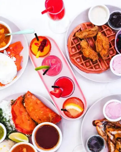 Colorful brunch flatlay — fried chicken and waffle, stuffed tacos, bowls of sauces and salsas, and vibrant fruit cocktails on a white table