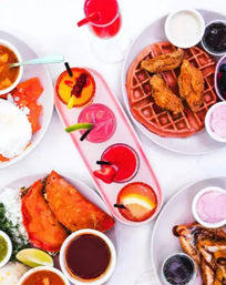 Colorful brunch flatlay — fried chicken and waffle, stuffed tacos, bowls of sauces and salsas, and vibrant fruit cocktails on a white table