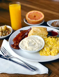 Classic American breakfast on a wooden table: fluffy scrambled eggs, crispy bacon, flaky biscuit with a bowl of gravy, small jam cup, glass of orange juice, halved grapefruit and a side of potatoes.
