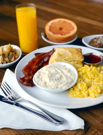 Classic American breakfast on a wooden table: fluffy scrambled eggs, crispy bacon, flaky biscuit with a bowl of gravy, small jam cup, glass of orange juice, halved grapefruit and a side of potatoes.