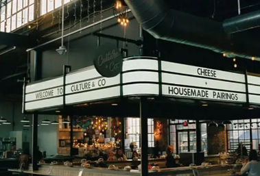 Artisan cheese counter in an industrial-style urban market hall with illuminated marquee reading 'CHEESE + HOUSEMADE PAIRINGS,' glass display cases, hanging pendant lights and large factory windows.