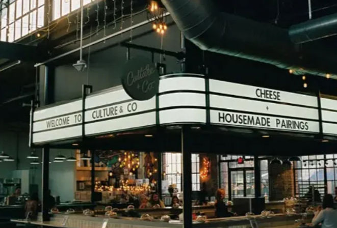 Artisan cheese counter in an industrial-style urban market hall with illuminated marquee reading 'CHEESE + HOUSEMADE PAIRINGS,' glass display cases, hanging pendant lights and large factory windows.