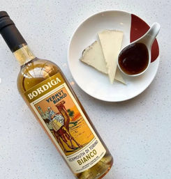 Bottle of white vermouth beside a plate with two wedges of soft cheese and a spoonful of dark fruit jam on a speckled kitchen countertop, aperitivo setup