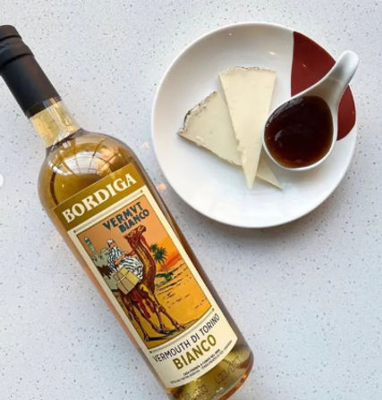 Bottle of white vermouth beside a plate with two wedges of soft cheese and a spoonful of dark fruit jam on a speckled kitchen countertop, aperitivo setup