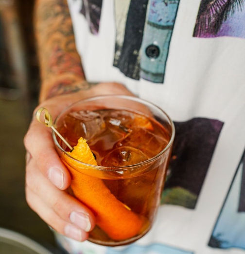 Hand with tattooed forearm holding a short glass of amber cocktail on the rocks, large ice cubes and an orange peel twist speared on a bamboo pick, colorful graphic t‑shirt in the background.