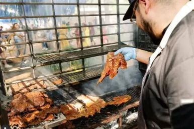 Smoky outdoor barbecue: gloved cook lifts a seasoned steak from a large open grill at a busy street-food market.