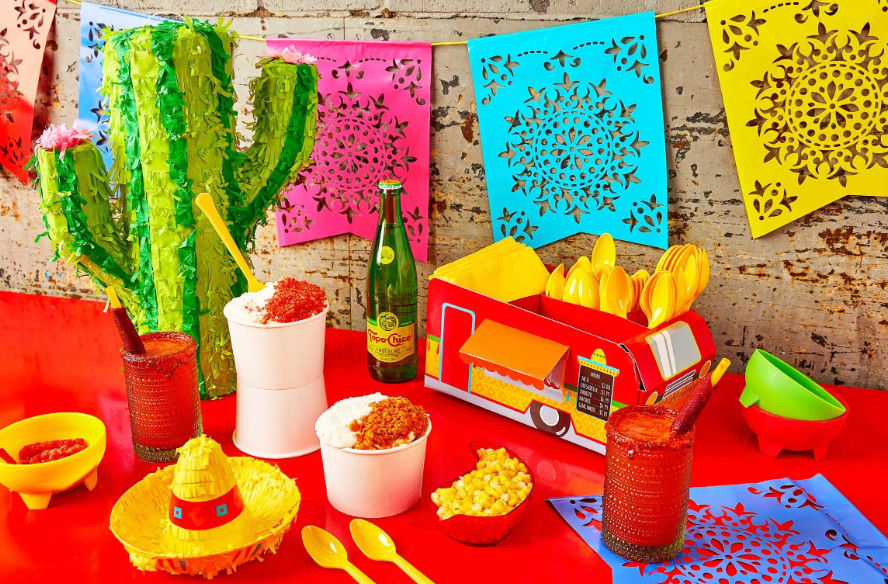 Vibrant Mexican fiesta table: papel picado banners, green cactus piñata, cardboard taco-truck utensil caddy, cups of chili-topped raspados, corn snack, red michelada drinks and yellow spoons on a red surface.