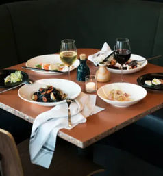 Cozy corner restaurant table with bowls of pasta and mussels, small salads, two glasses of red and white wine, olive oil bottle, candle and a draped napkin.
