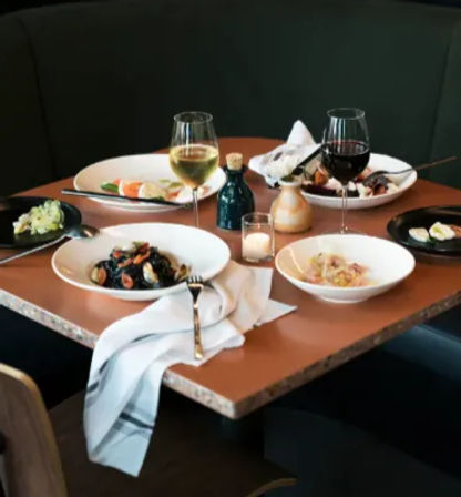 Cozy corner restaurant table with bowls of pasta and mussels, small salads, two glasses of red and white wine, olive oil bottle, candle and a draped napkin.
