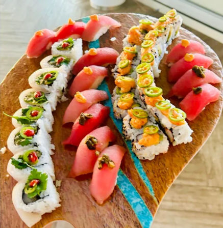 Assorted sushi platter with fresh tuna nigiri and two types of maki rolls—one topped with jalapeño slices and orange sauce, another with leafy greens and red chili—arranged on a wooden serving board with a turquoise resin inlay.