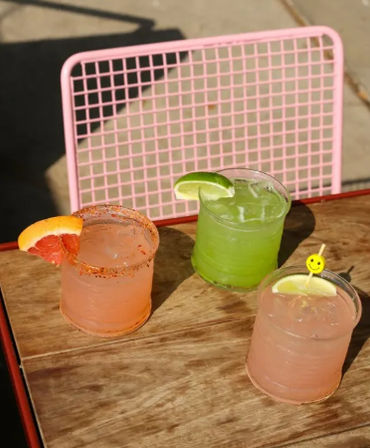 Three colorful summer cocktails on a wooden patio table: a pink grapefruit drink with chili-rim and grapefruit wedge, a bright green lime cocktail with lime wedge, and a pale pink drink with lemon slice and smiley stirrer, set against a pink wire patio chair.