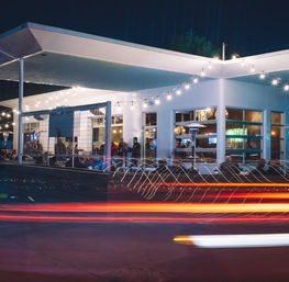 Urban nighttime restaurant patio with string lights and outdoor diners at a modern white building, long-exposure red and white car light trails streaking past.
