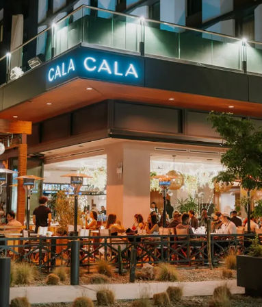 Evening city patio dining at a modern corner restaurant — bustling outdoor seating with warm string lights, patio heaters, neon signage, and groups enjoying meals