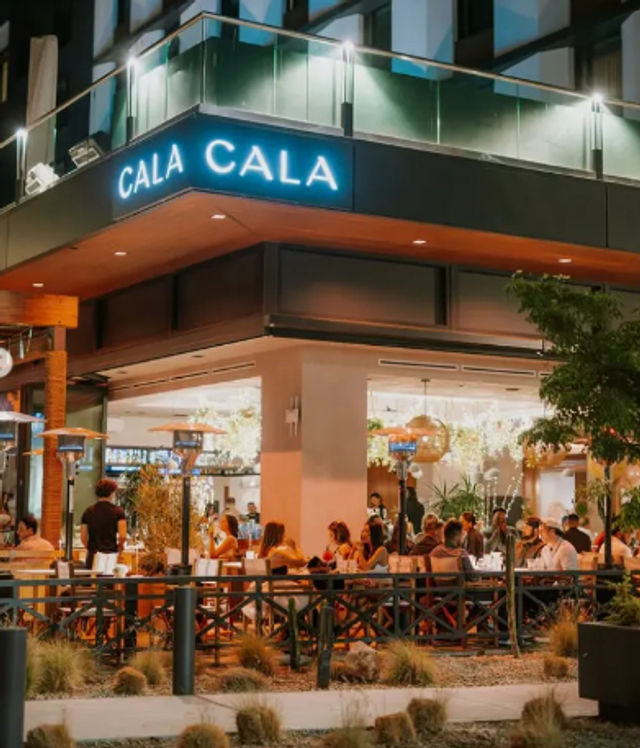 Evening city patio dining at a modern corner restaurant — bustling outdoor seating with warm string lights, patio heaters, neon signage, and groups enjoying meals