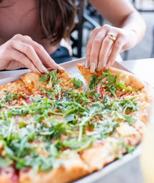 Hands grabbing a slice of arugula-topped thin-crust pizza sprinkled with grated cheese at an outdoor cafe