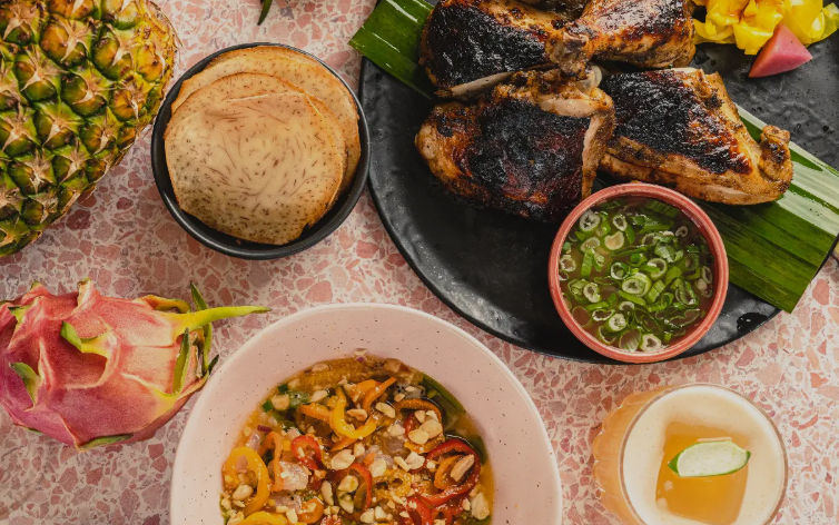 Vibrant tropical spread: charred grilled chicken on a banana leaf with scallion dipping sauce, taro chips, a colorful pepper-and-nut salad, fresh pineapple and dragon fruit, and a tropical cocktail.