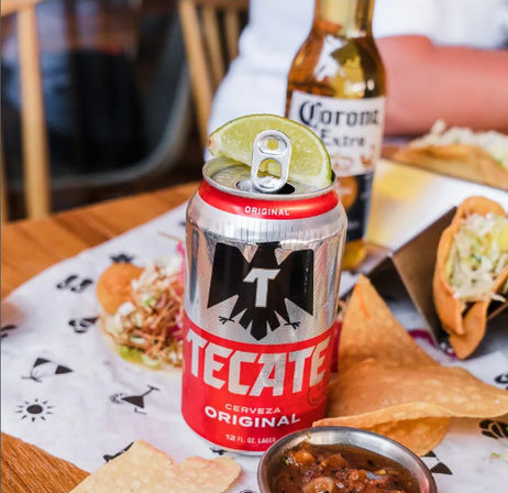 Tecate beer can with lime wedge on a table surrounded by tacos, tortilla chips and salsa, casual Mexican-style meal with a Corona bottle blurred in the background