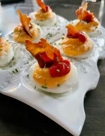 Bite-sized deviled eggs crowned with crispy bacon shards, bright red pepper sauce and chopped chives on a white ceramic serving platter