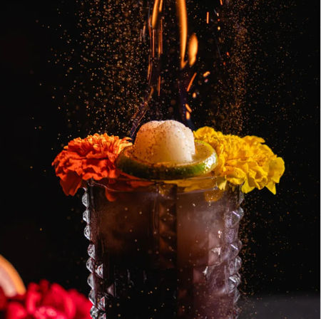 Smoky cocktail in a textured glass topped with an ice sphere on a lime slice, garnished with orange and yellow marigold flowers and flaming sparks against a dark background.