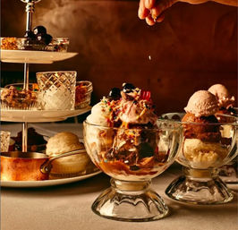 Glass dessert bowls with decadent ice cream sundaes — scoops of vanilla, chocolate and strawberry topped with cherries, sprinkles and caramel drizzle while a hand adds a finishing garnish, with a tiered tray of pastries and sauces in the background.