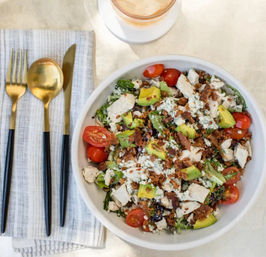 Top-down view of a chicken and avocado salad with cherry tomatoes, blue cheese crumbles and bacon bits in a white bowl, gold-and-black cutlery on a striped napkin and an iced drink nearby
