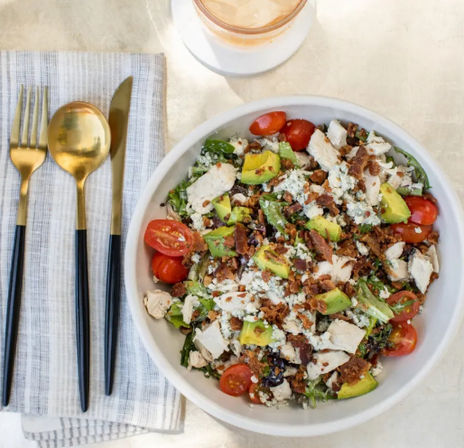 Top-down view of a chicken and avocado salad with cherry tomatoes, blue cheese crumbles and bacon bits in a white bowl, gold-and-black cutlery on a striped napkin and an iced drink nearby