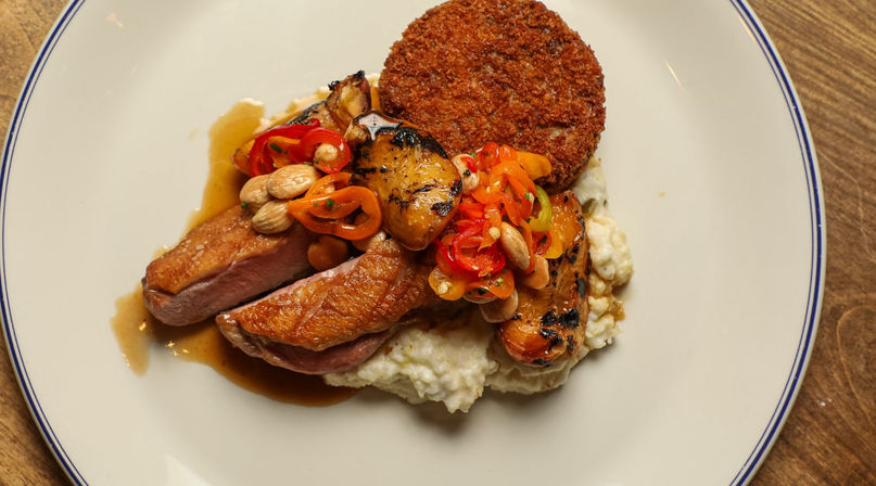 Restaurant-style seared duck breast on creamy mashed potatoes topped with grilled apricot, pickled peppers, toasted almonds, pan sauce and a crispy potato cake on a white plate.
